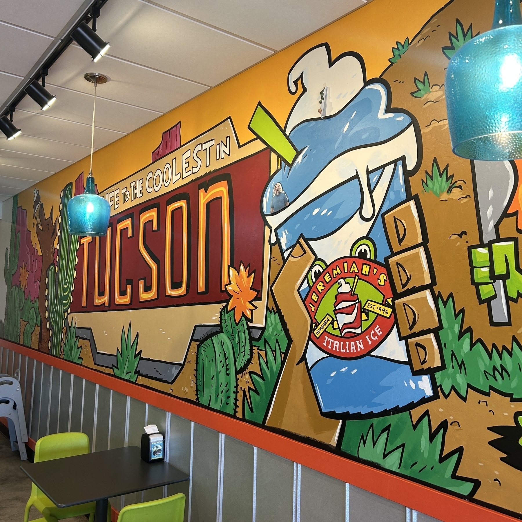 A vibrant mural featuring a retro sign reading "Voted the Coolest in TUCSON" alongside colorful artwork of a person holding an Italian Ice.
