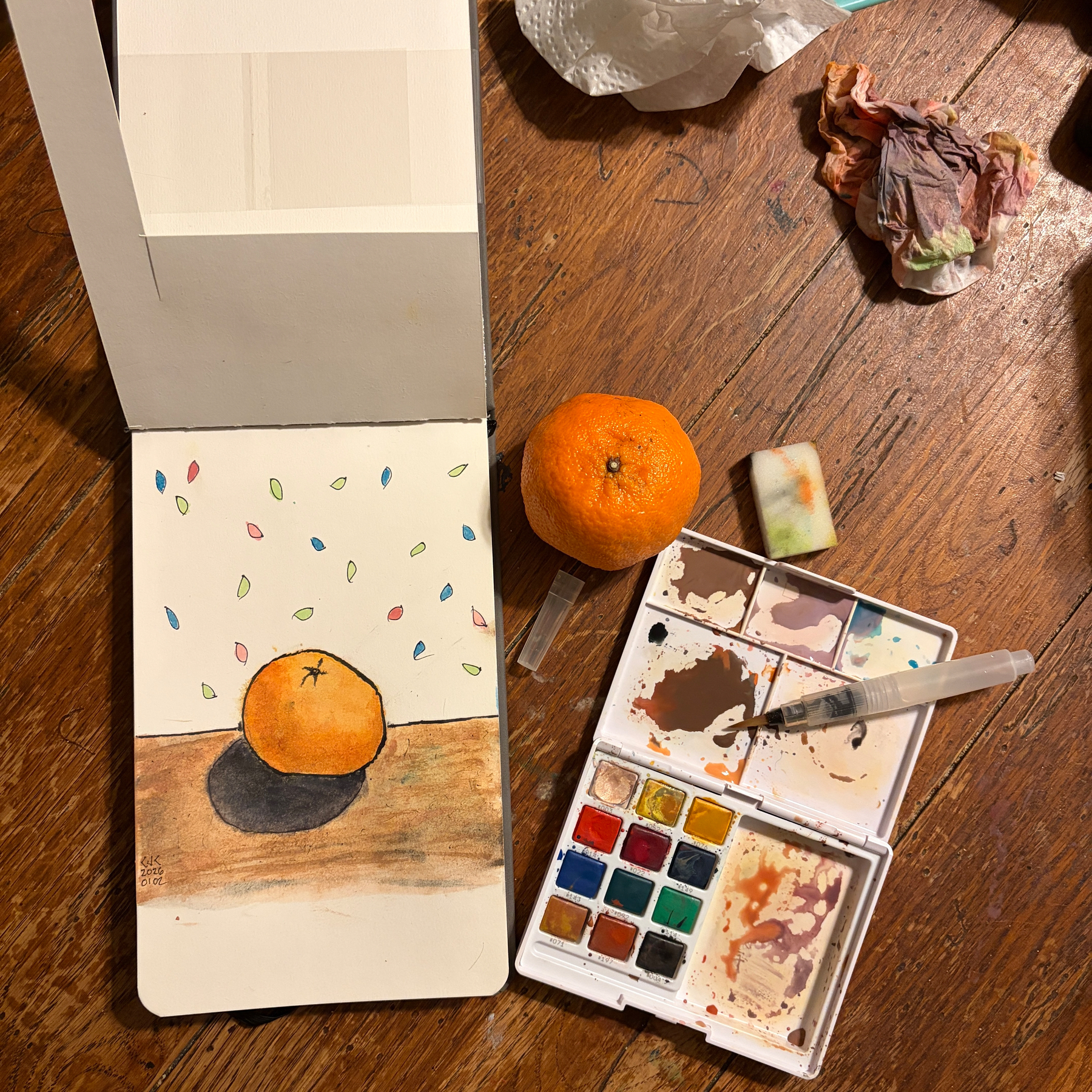 A watercolor painting of an orange sits next to a real orange, a partially used paint palette, and crumpled paper towels on a wood table.
