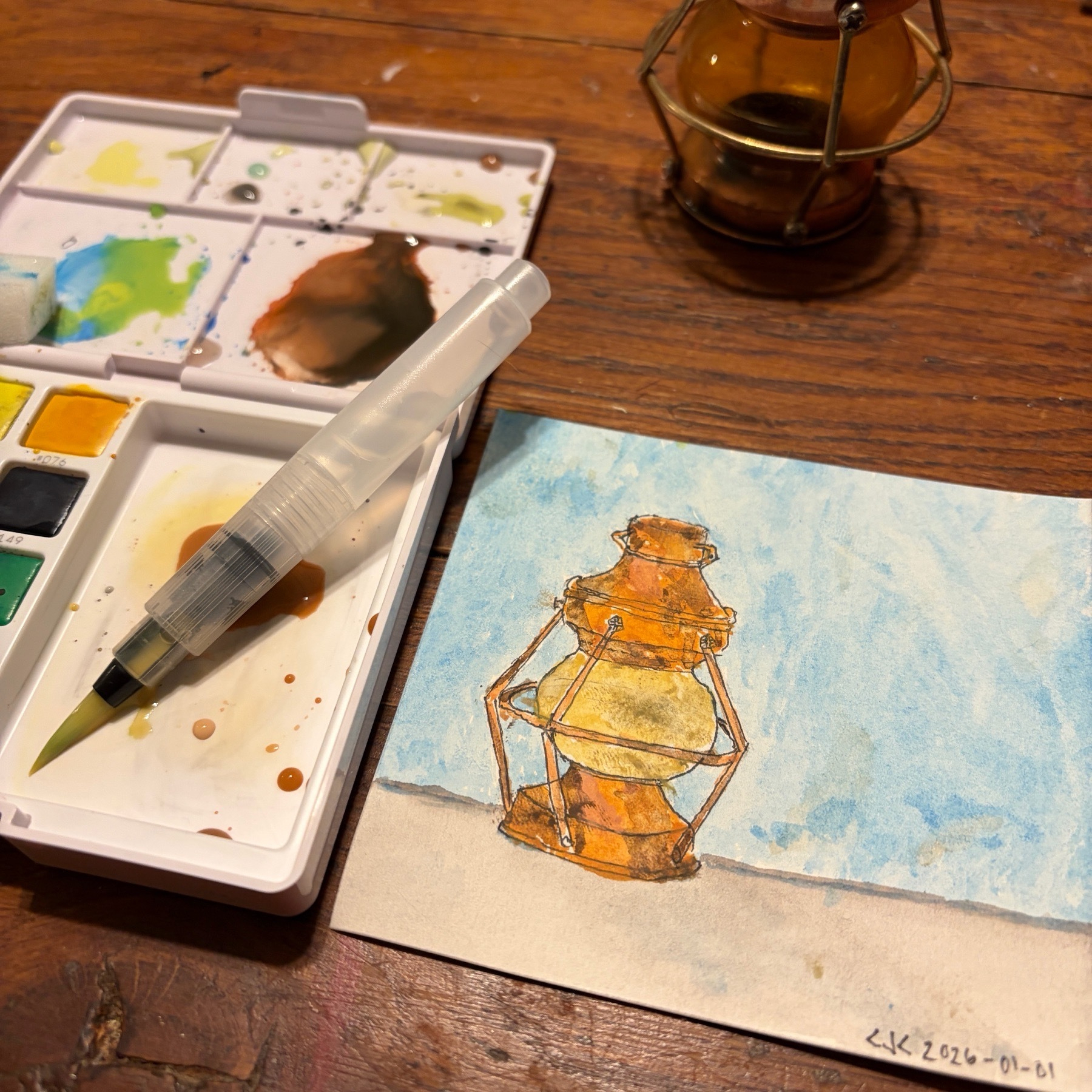Watercolor painting of a latern with the latent nearby and an artists watercolor palette and brush with the colors mixed as used in the painting.