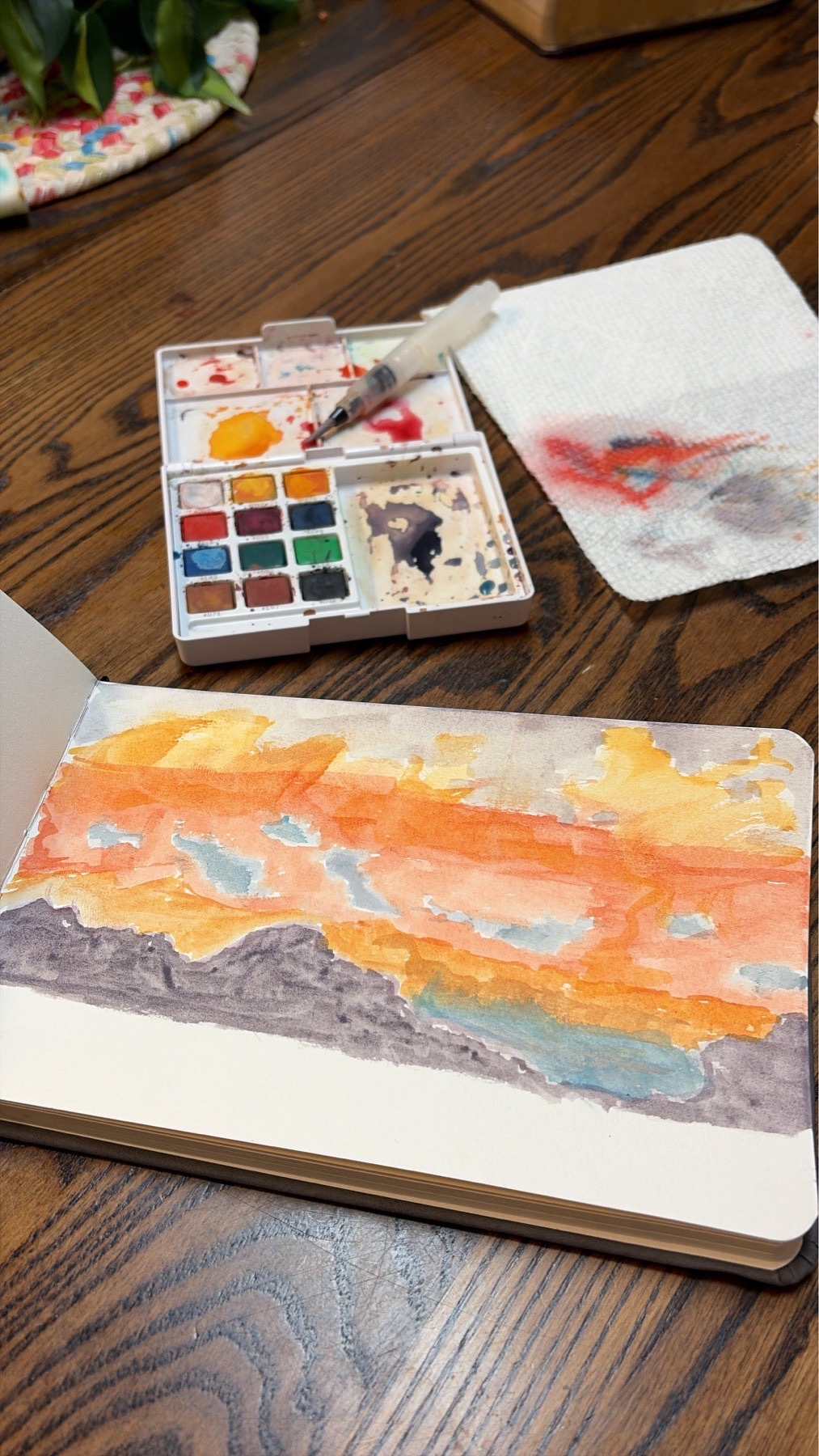 Watercolor painting of a sunset with the used paints and brush nearby.